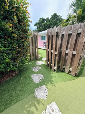 A garden pathway with a wooden fence, green artificial grass, and a pink building in the background.