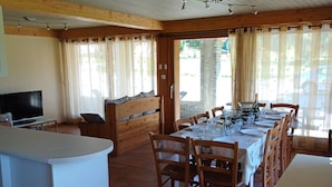 Un comedor con una mesa larga dispuesta para seis personas, sillas de madera, una encimera blanca y un televisor sobre una mesita.