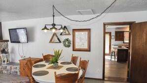 A dining area with a round table, wooden chairs, a potted plant, and a television mounted on the wall. - River Rider at Brookside (Red River)