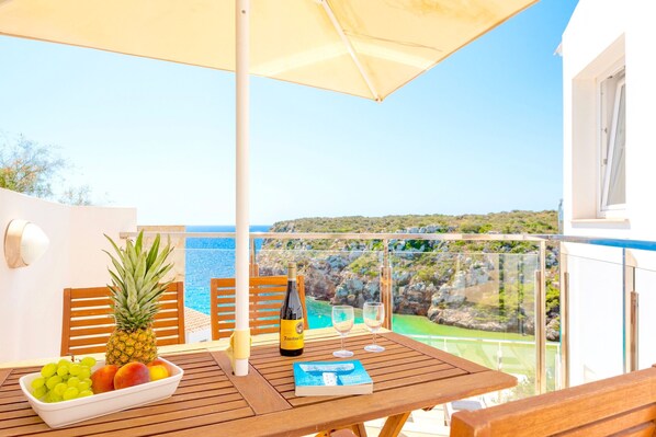 A wooden table with a bowl of fruit, a bottle of wine, and a book on a balcony overlooking a scenic view. - Seafront Villa Bellavista with private heated pool (Calan Porter)