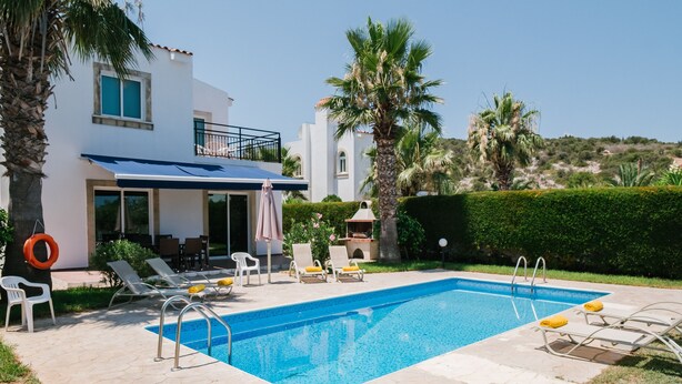 A pool area with lounge chairs and a lifebuoy, surrounded by palm trees and a building with a balcony. - Chrysanthemum Villa (Neo Chorio)