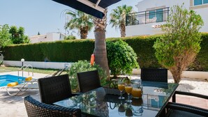 An outdoor dining area with a glass table, wicker chairs, and a pool in the background. - Chrysanthemum Villa (Neo Chorio)
