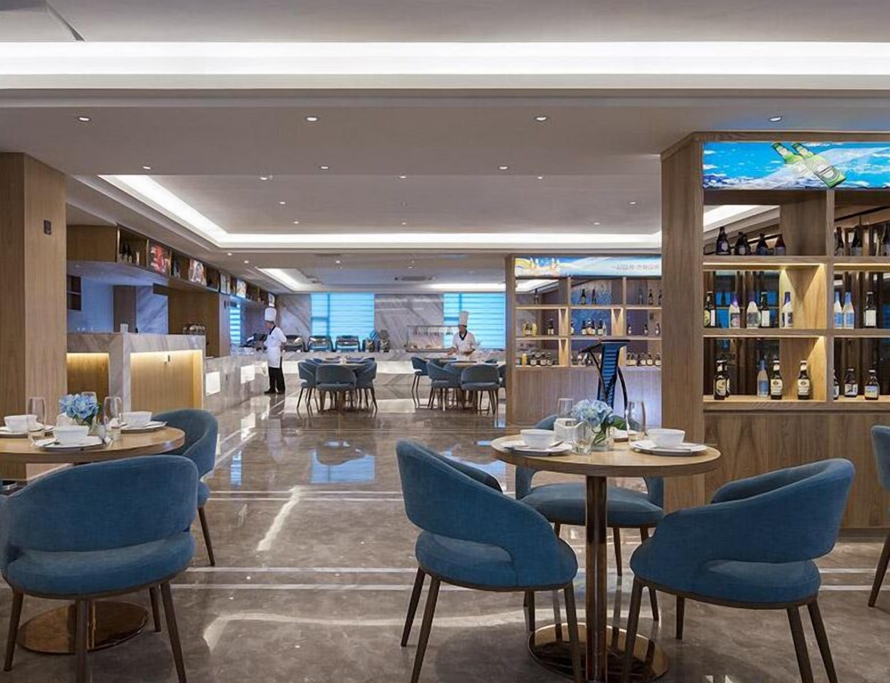A modern restaurant with round tables and blue chairs, a bar area with shelves of bottles, and a chef preparing food.