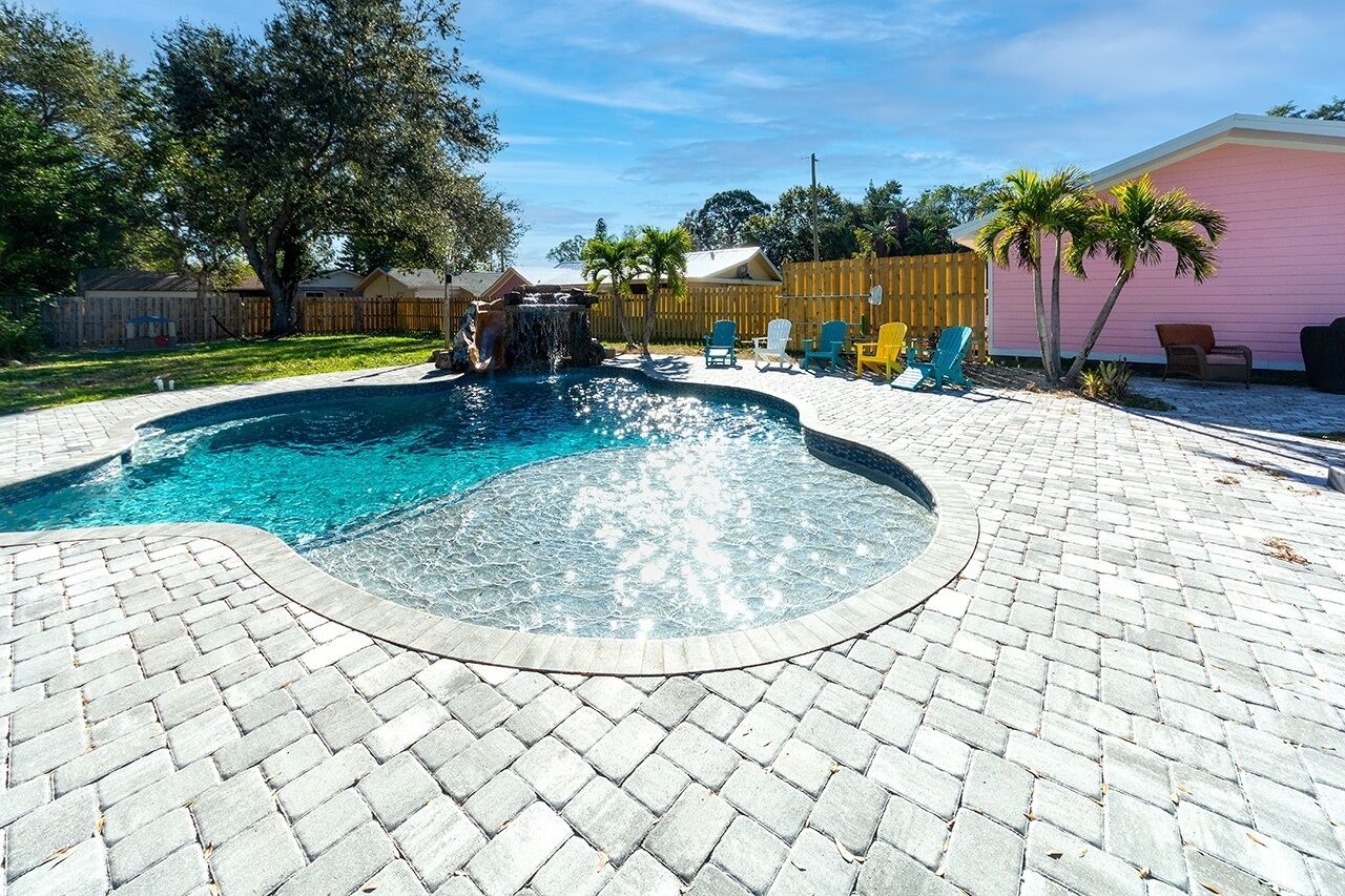A swimming pool with a waterfall feature, surrounded by a paved patio with chairs and a wooden fence.