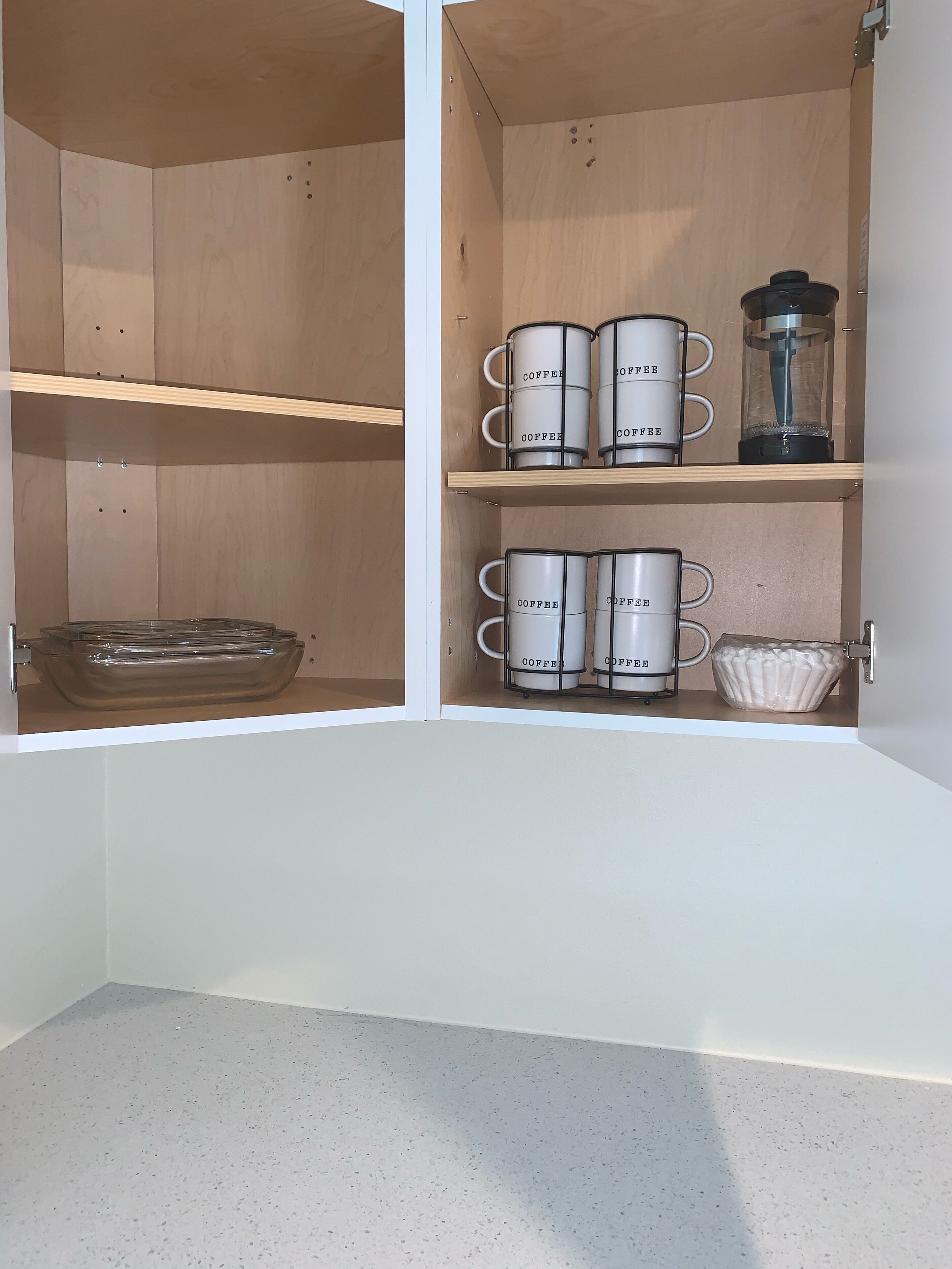 A kitchen cabinet with two shelves, each holding multiple white mugs with the word 'COFFEE' on them, a glass coffee pot, and a clear glass bowl.