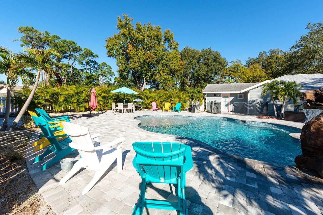 En swimmingpool med en stenbelagd poolkant, omgiven av färgglada Adirondack-stolar och parasoller, med träd i bakgrunden.