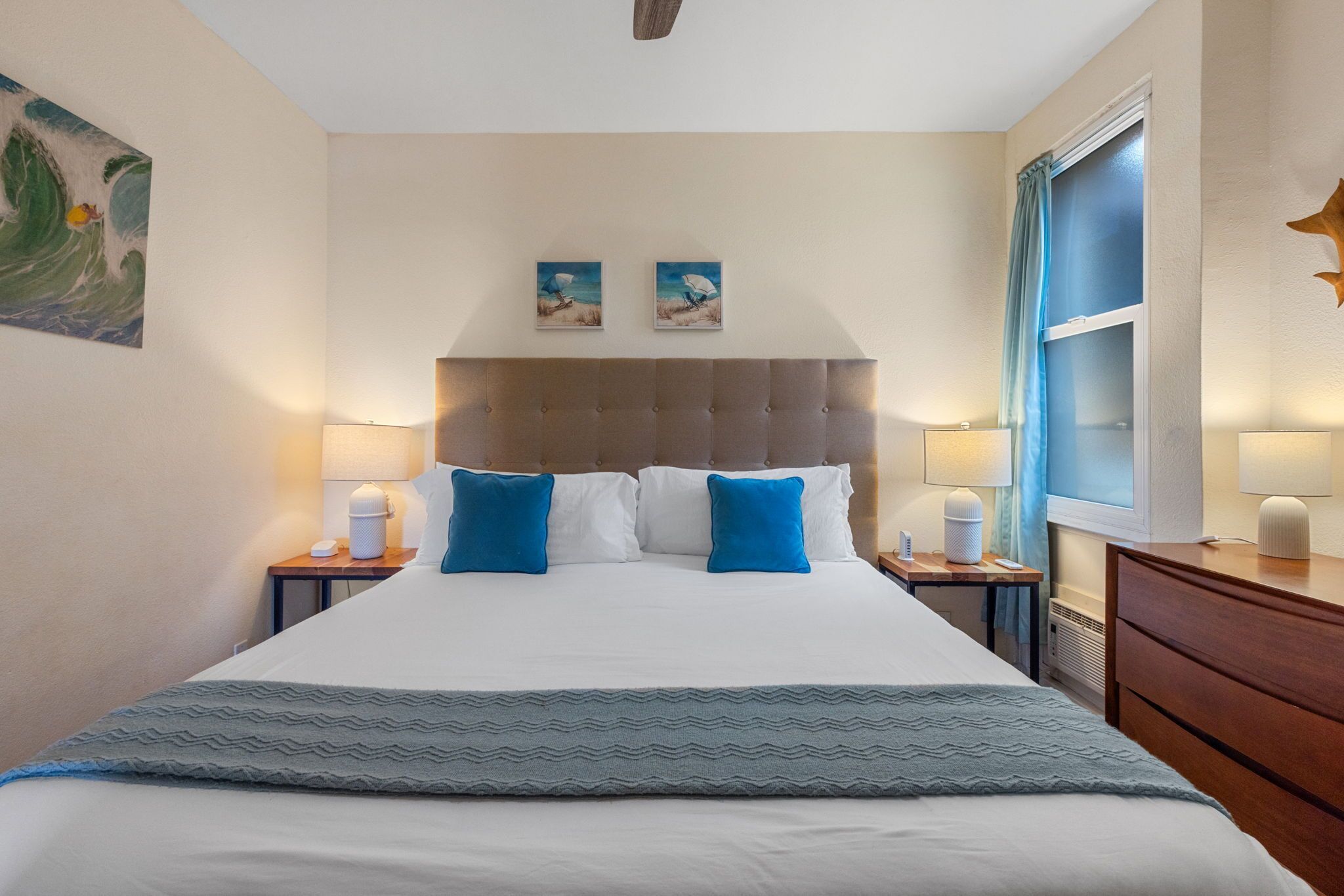 A hotel room with a large bed, two bedside tables, a nightstand, a dresser, a window with curtains, and two wall-mounted paintings.