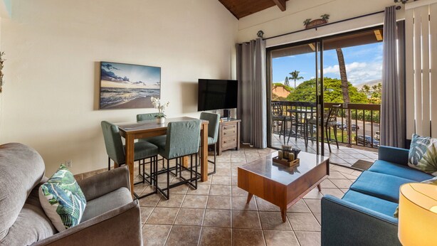 A spacious living area with a dining table, chairs, a sofa, a coffee table, and a TV. There is a balcony with a view of the outdoors. - Beautiful, Family Friendly 2 Br 2Ba Across Street From Beach. (Kihei)