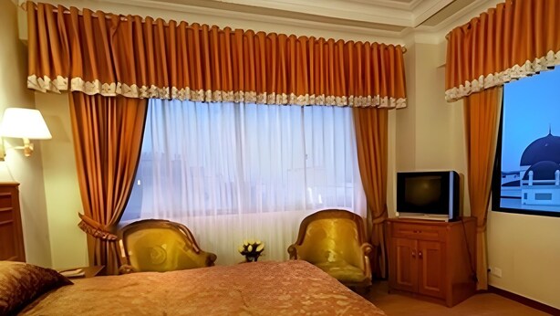 Una habitación de hotel con una cama grande, dos sillones, un televisor y un ventanal con cortinas.