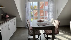 Spiseområde med bord og stole, et vindue med gardiner, samt et køkken med skabe og elkedel.
