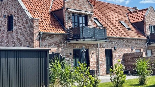Et toetages murhus med rødt tegltag, en balkon og et lille haveområde.