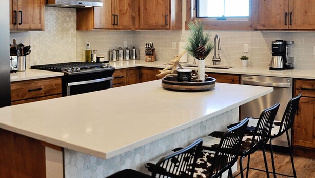 A modern kitchen with wooden cabinets, a large island, bar stools, pendant lighting, and a window providing natural light. - BRAND NEW Contemporary 2 BR near Winter Park Ski Resort on Shuttle w/priv hottub (fraser)