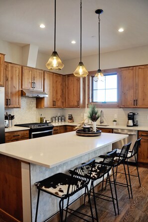 A modern kitchen with wooden cabinets, a large island, bar stools, pendant lighting, and a window providing natural light. - BRAND NEW Contemporary 2 BR near Winter Park Ski Resort on Shuttle w/priv hottub (fraser)