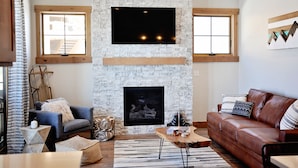 A living room with a stone fireplace, two windows, a brown leather sofa, and a grey armchair. - BRAND NEW Contemporary 2 BR near Winter Park Ski Resort on Shuttle w/priv hottub (fraser)