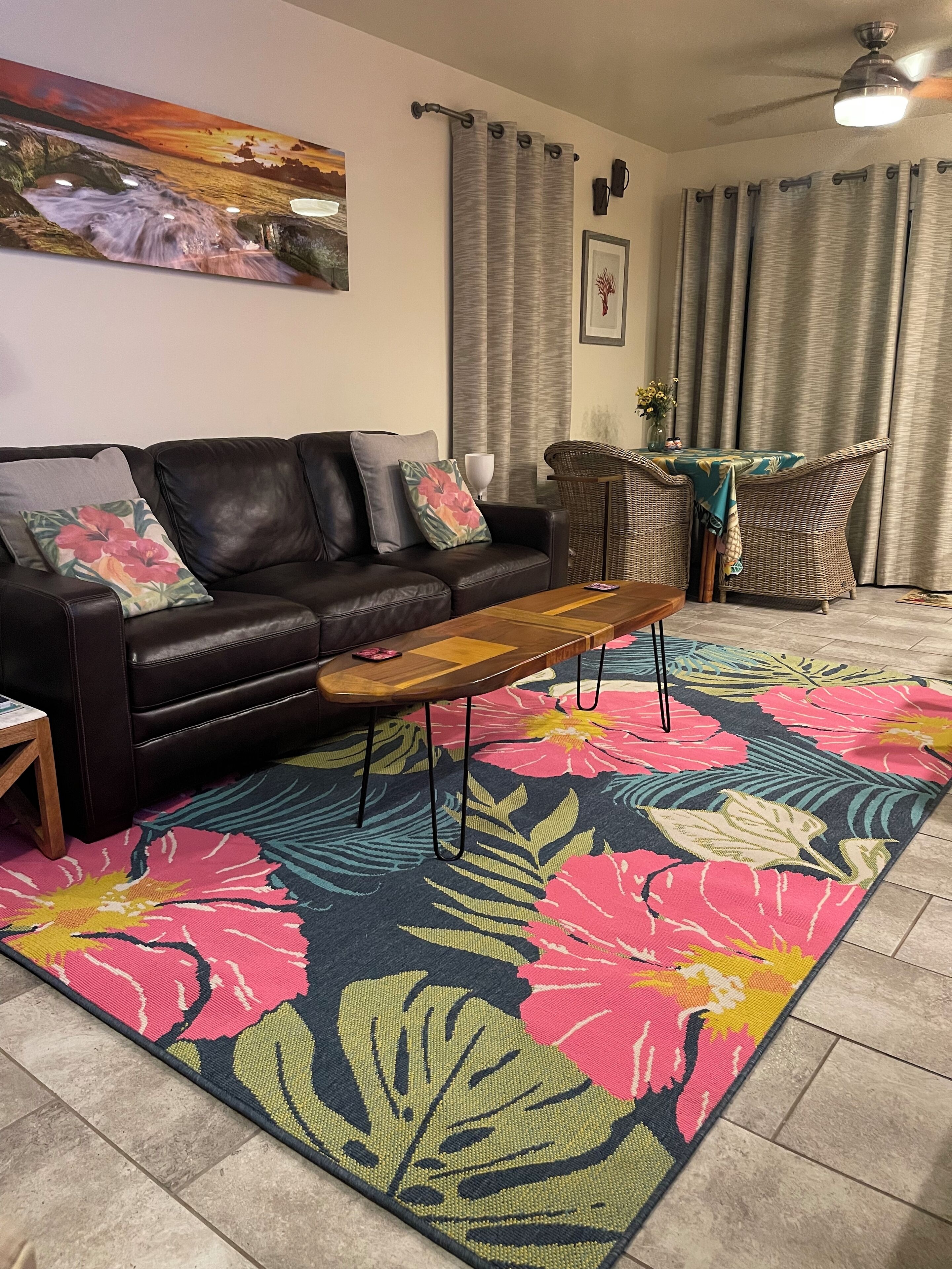 A living room with a black leather sofa, a wooden coffee table, a colorful floral rug, a wicker chair, and a ceiling fan.