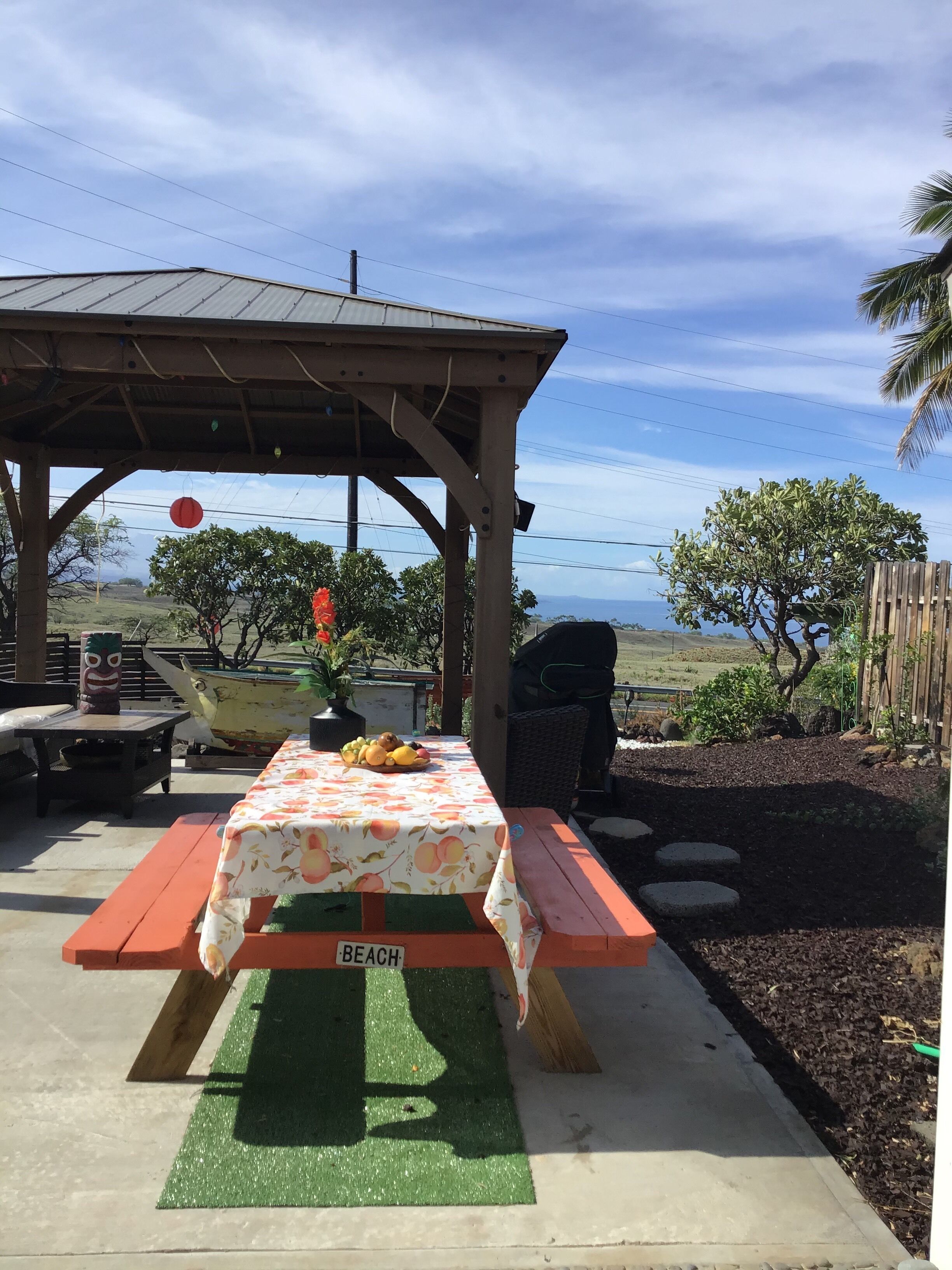 Ein Picknicktisch mit einem 'BEACH'-Schild, umgeben von einer Holzpergola, einer Tiki-Statue und einem Boot.