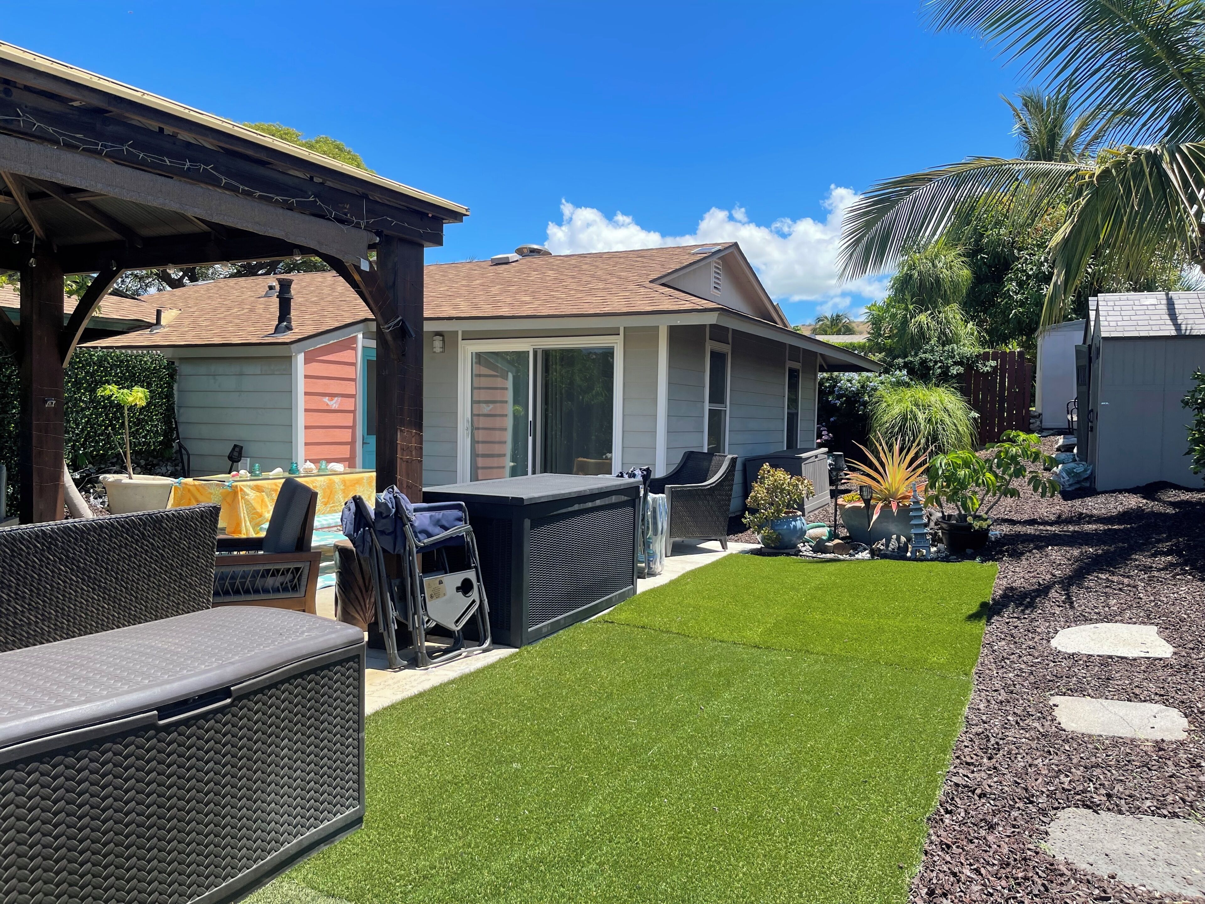 Un jardin avec une pelouse artificielle, du mobilier d’extérieur, un patio couvert et une maison à l’extérieur rouge et aux bordures blanches.