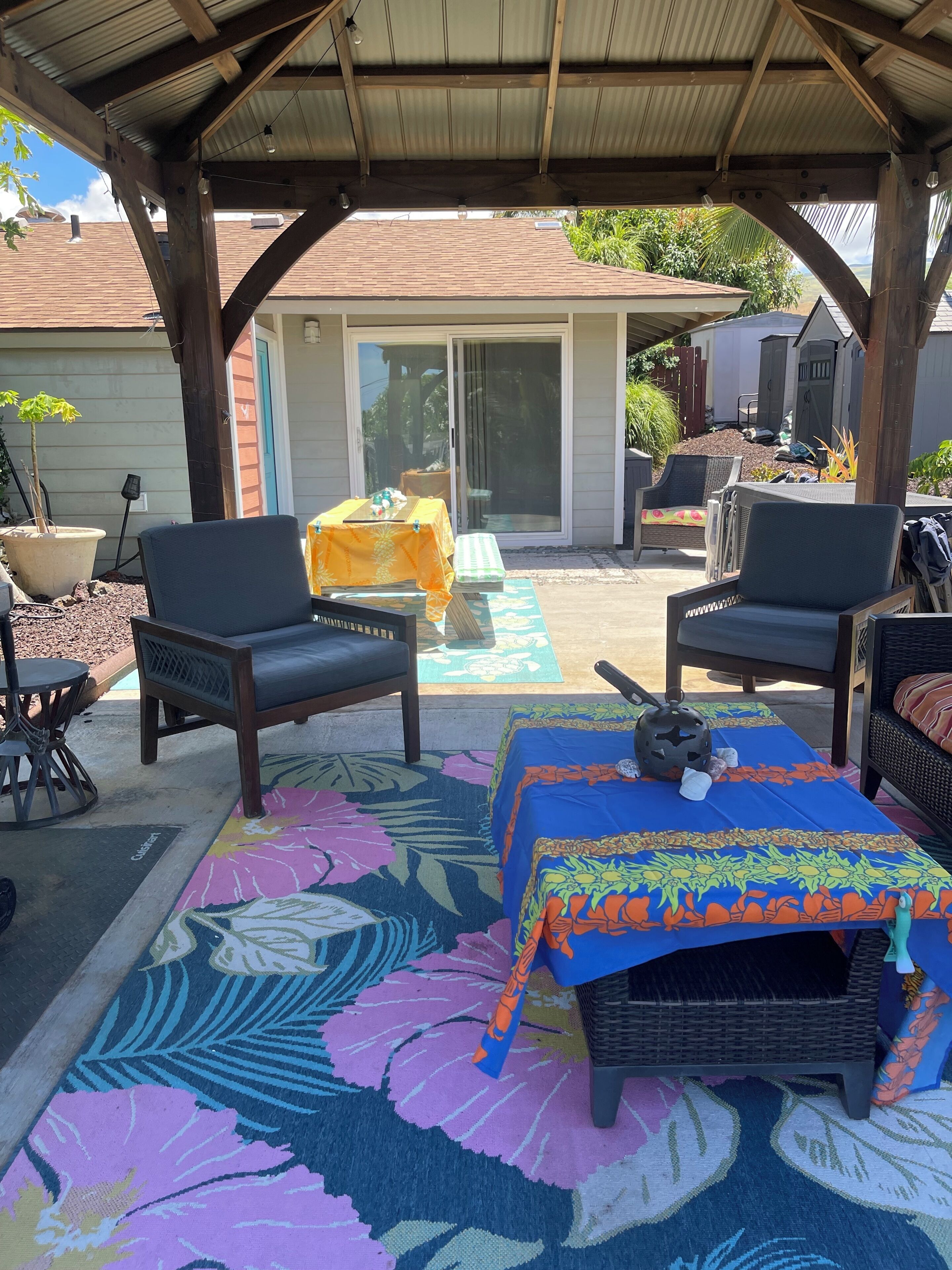 A covered patio area with outdoor furniture, a table with a yellow tablecloth, and a colorful rug.