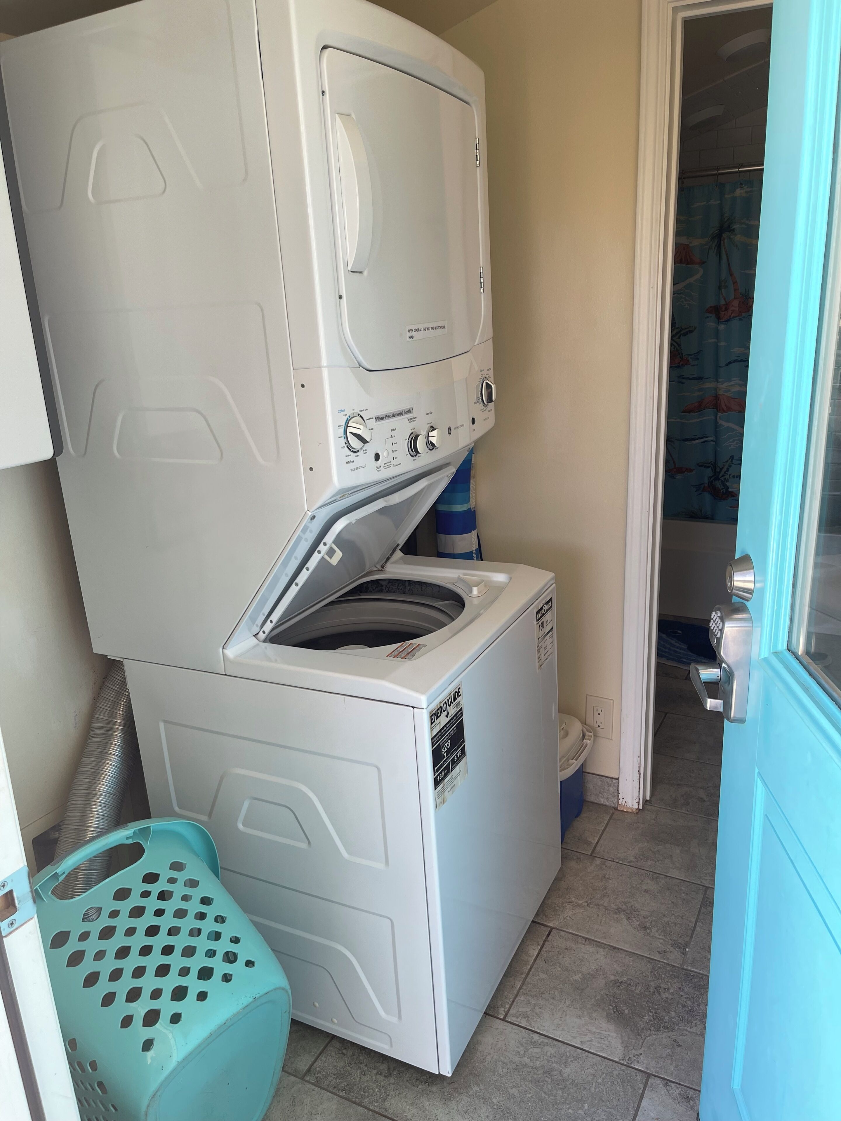 Un lavadero pequeño con lavadora y secadora de carga frontal, una cesta de ropa azul y una puerta que da a otra habitación.