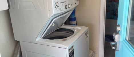 Un lavadero pequeño con lavadora y secadora de carga frontal, una cesta de ropa azul y una puerta que da a otra habitación.