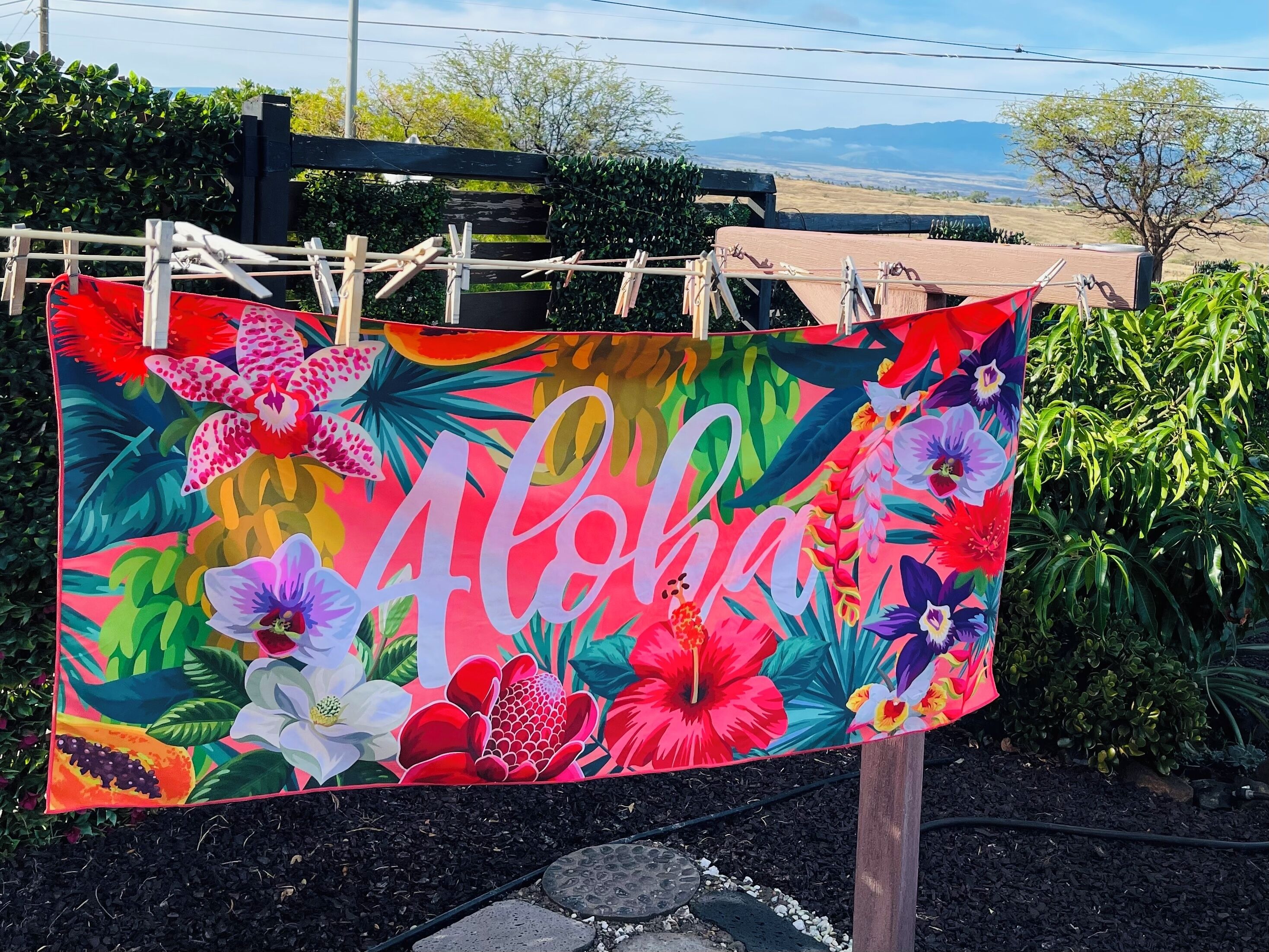 Una toalla colorida con flores tropicales y la palabra “Aloha” estampada, colgada en una cerca de madera.