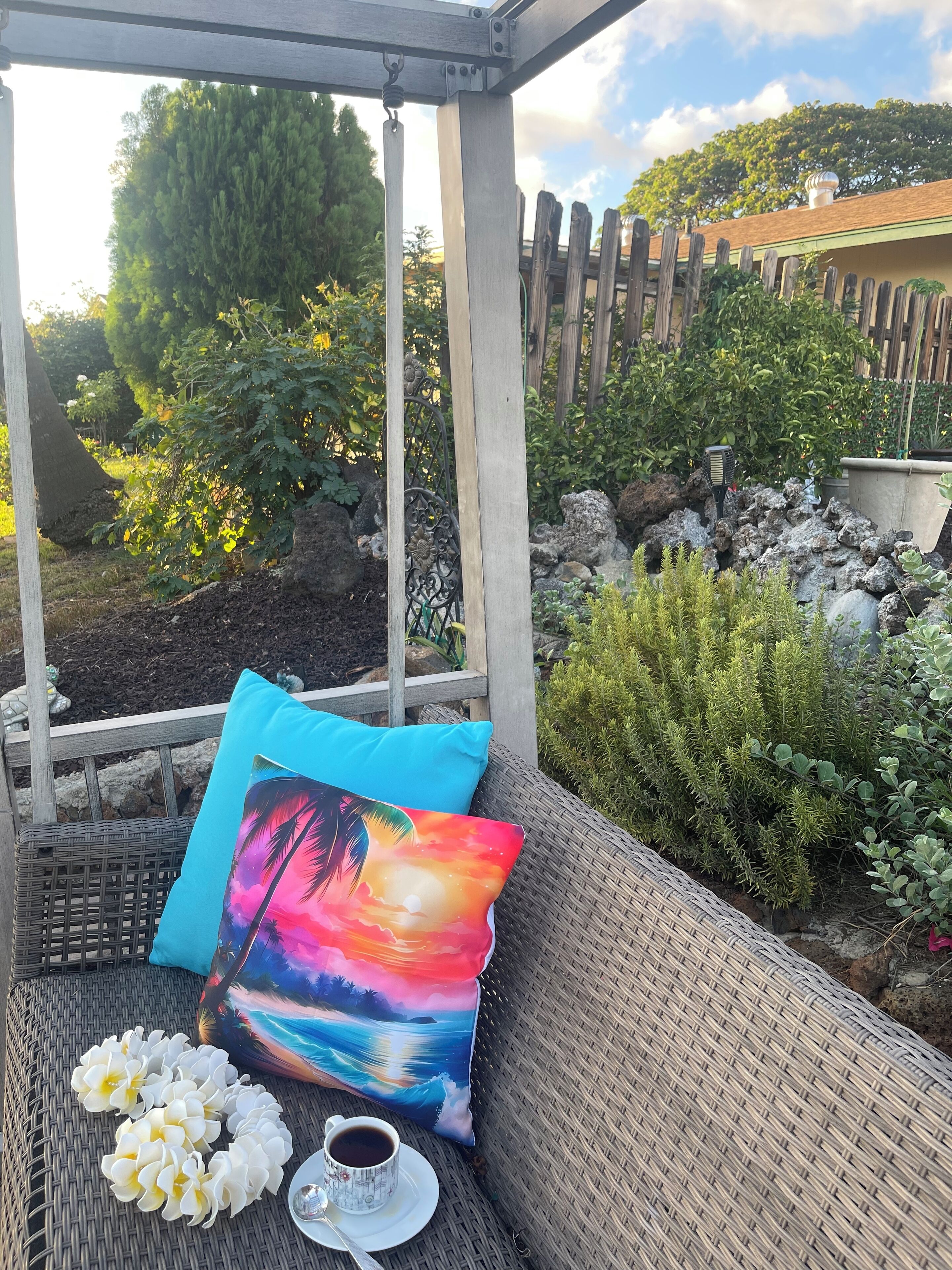 Ein überdachter Außenbereich mit Rattan-Sofa, einem bunten Kissen, weißen Blumen und einer Kaffeetasse.