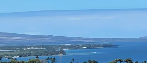Paisatge costaner amb vista clara a l'oceà, una línia de costa llunyana amb edificis, i un primer pla de terreny rocós i sec.
