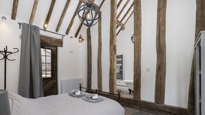 Ein Schlafzimmer mit freiliegenden Holzbalken, einem Bett mit grauer Bettwäsche, einer Pendelleuchte und Blick auf ein Badezimmer durch eine Holztür.