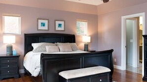 Quarto com cama de madeira escura, duas luminárias de cabeceira, um ventilador de teto e um banco branco.