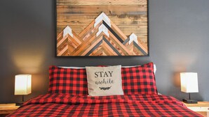 A bed with a red and black checkered bedspread, a pillow with the text "STAY awhile," and a wooden wall art piece above the bed. - Mountain/River Rider at Brookside (Red River)