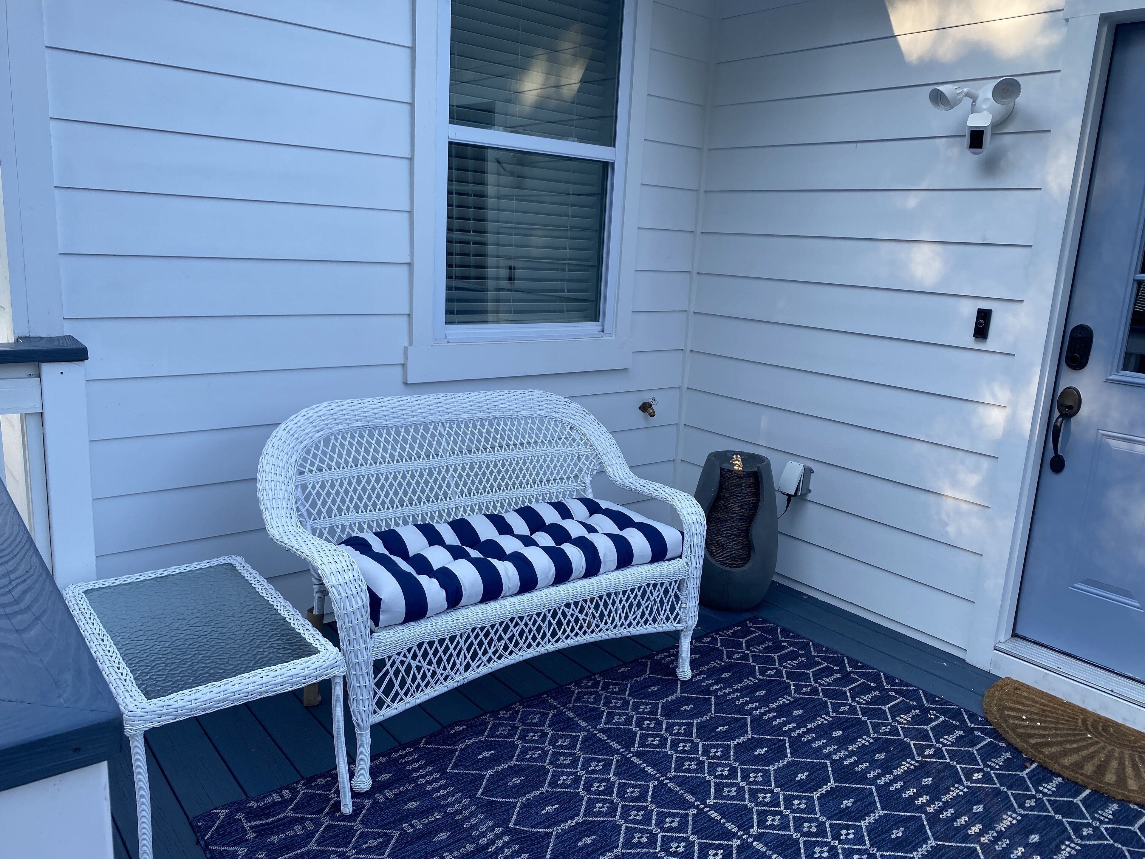 A white wicker chair with a striped cushion on a dark patterned rug, a small white table, and a door with a security camera.