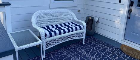 A white wicker chair with a striped cushion on a dark patterned rug, a small white table, and a door with a security camera.
