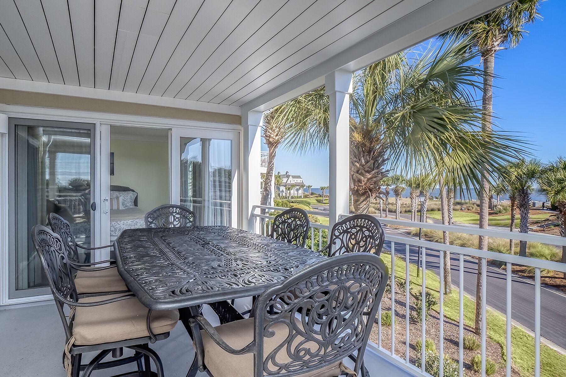 A covered outdoor patio with a table and chairs, a view of palm trees, and a road in the background.