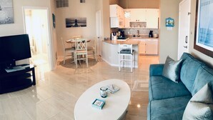 A living room with a blue sofa, a coffee table, and a TV. There is a kitchen in the background with white cabinets and a dining area with a table and chairs. - "The Dover" ~ a glimpse of the sea Resort-style condo (Corpus Christi)