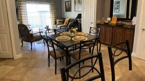 Un comedor con una mesa preparada para cuatro personas, rodeada de sillas. Se observa una cocina al fondo con una encimera y armarios.