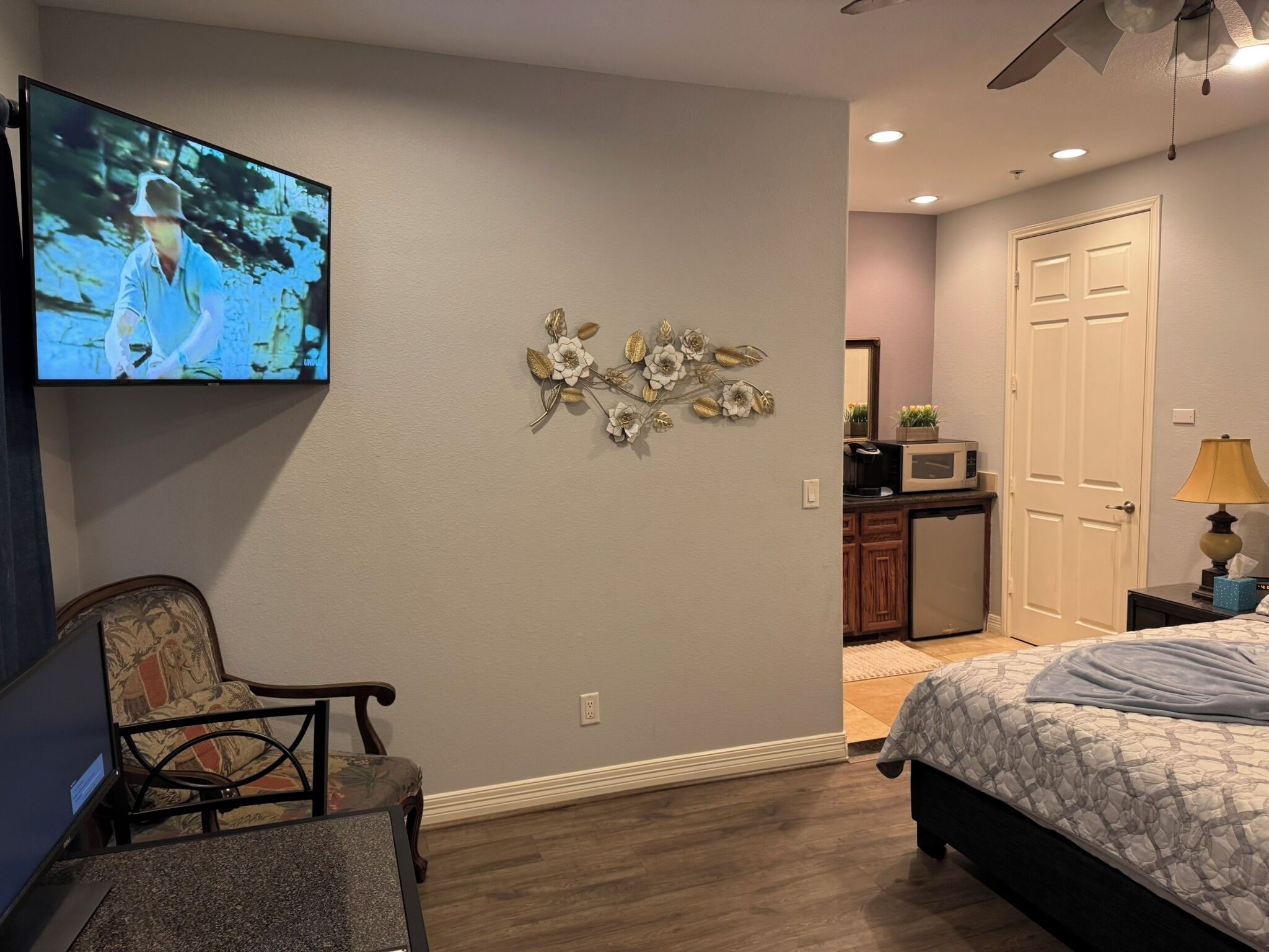 A bedroom with a bed, a TV mounted on the wall, a decorative wall piece, a chair, and a desk with a microwave.