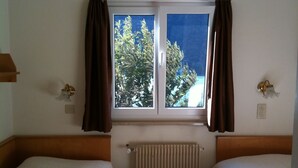 A hotel room with two single beds, a window with curtains, a radiator, and a view of greenery outside.