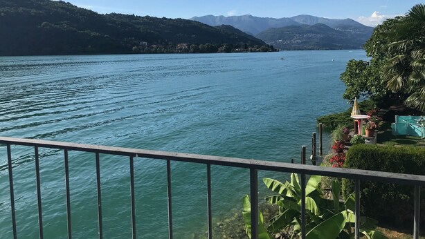 A lake with mountains in the background, a railing in the foreground, and some greenery on the right side.