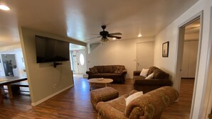 A spacious living room with hardwood flooring, a ceiling fan, and a flat-screen TV mounted on the wall.