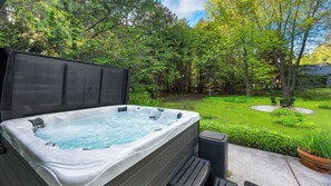 A hot tub in a lush garden with a wooden deck and a trash can nearby.
