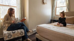 A room with a bed, a window, a chair, and a person reading a book.