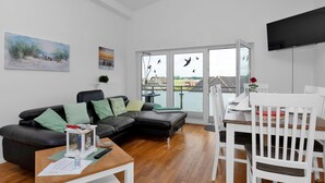 A modern living room with a black leather sofa, white dining table, and chairs. There is a large window with a view of the outside. - Apartment Sturm by Interhome (Warwerort)