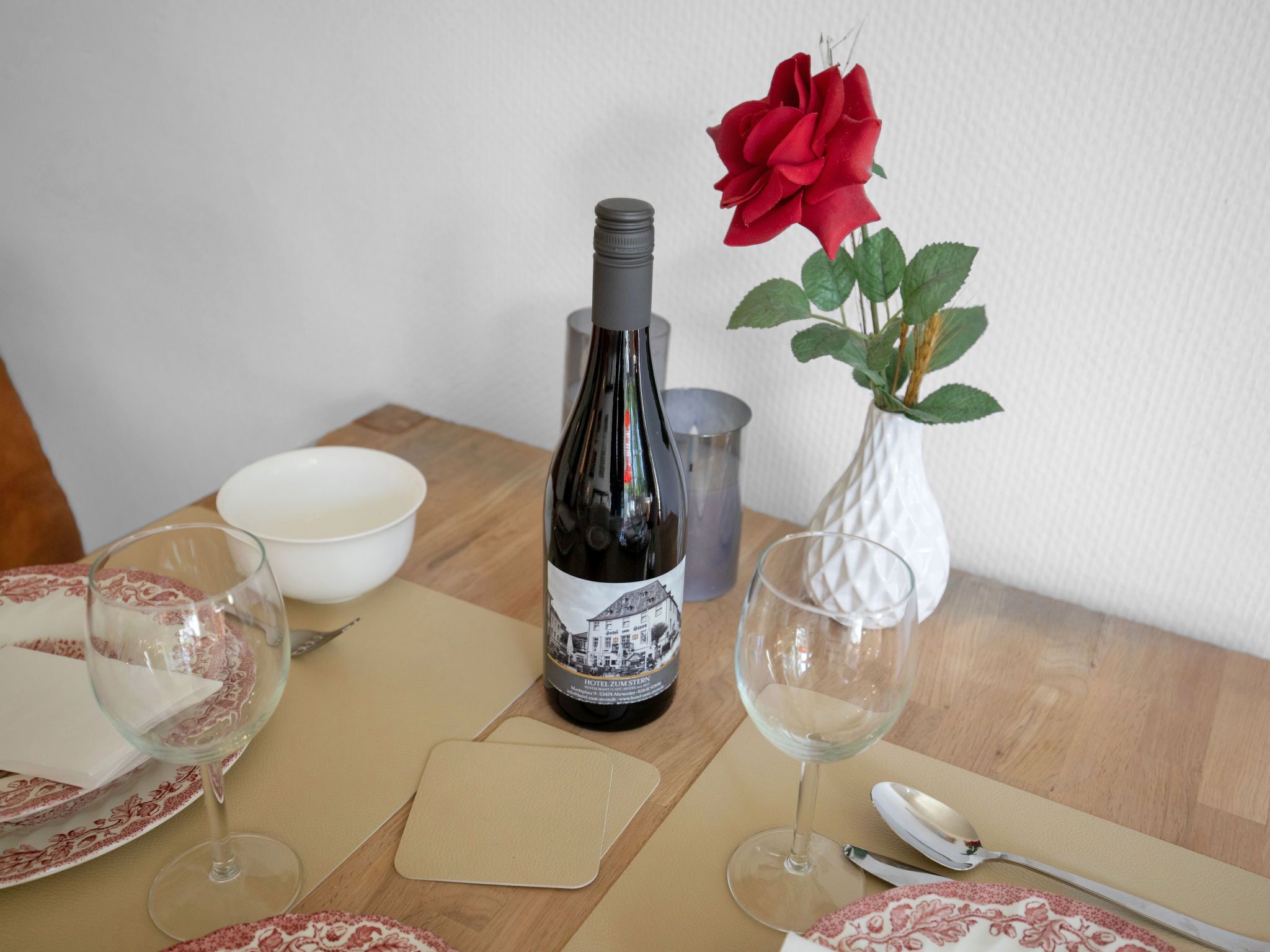Une table à manger avec une bouteille de vin, un vase contenant une rose rouge et des verres à vin vides.