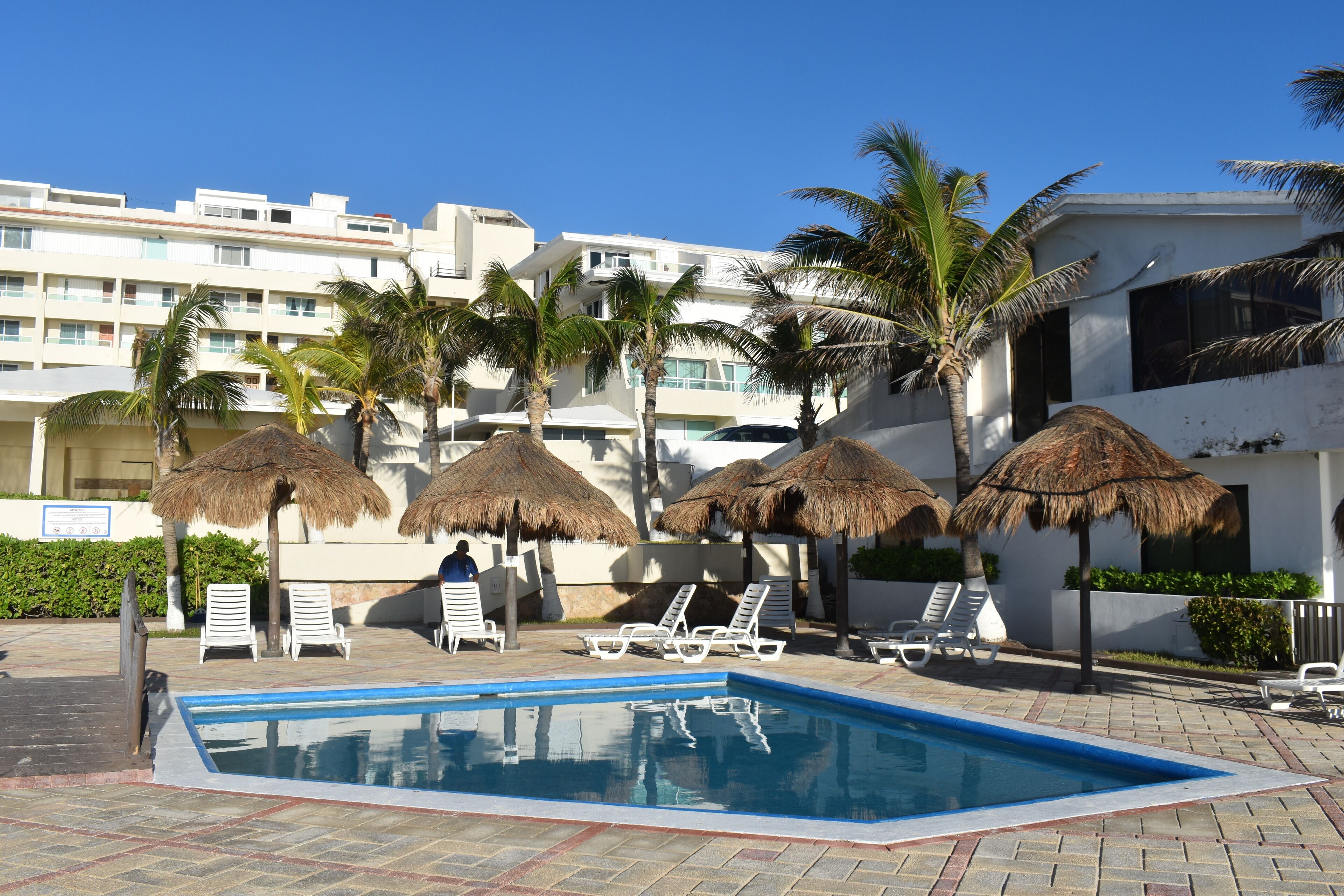 A swimming pool with lounge chairs and thatched umbrellas, surrounded by palm trees and a white building.