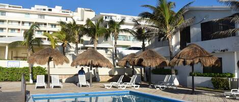 A swimming pool with lounge chairs and thatched umbrellas, surrounded by palm trees and a white building.