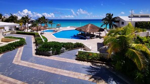 A resort pool with a view of the ocean, surrounded by palm trees and thatched-roof structures. - Cozy Studio In Beachfront Condominium (Cancun)