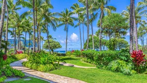 A tropical garden with palm trees, a stone pathway, and a view of the ocean. - Turtle Bay Villas 108 by Gather w/ Shared Pool, Spa, AC (Kahuku)