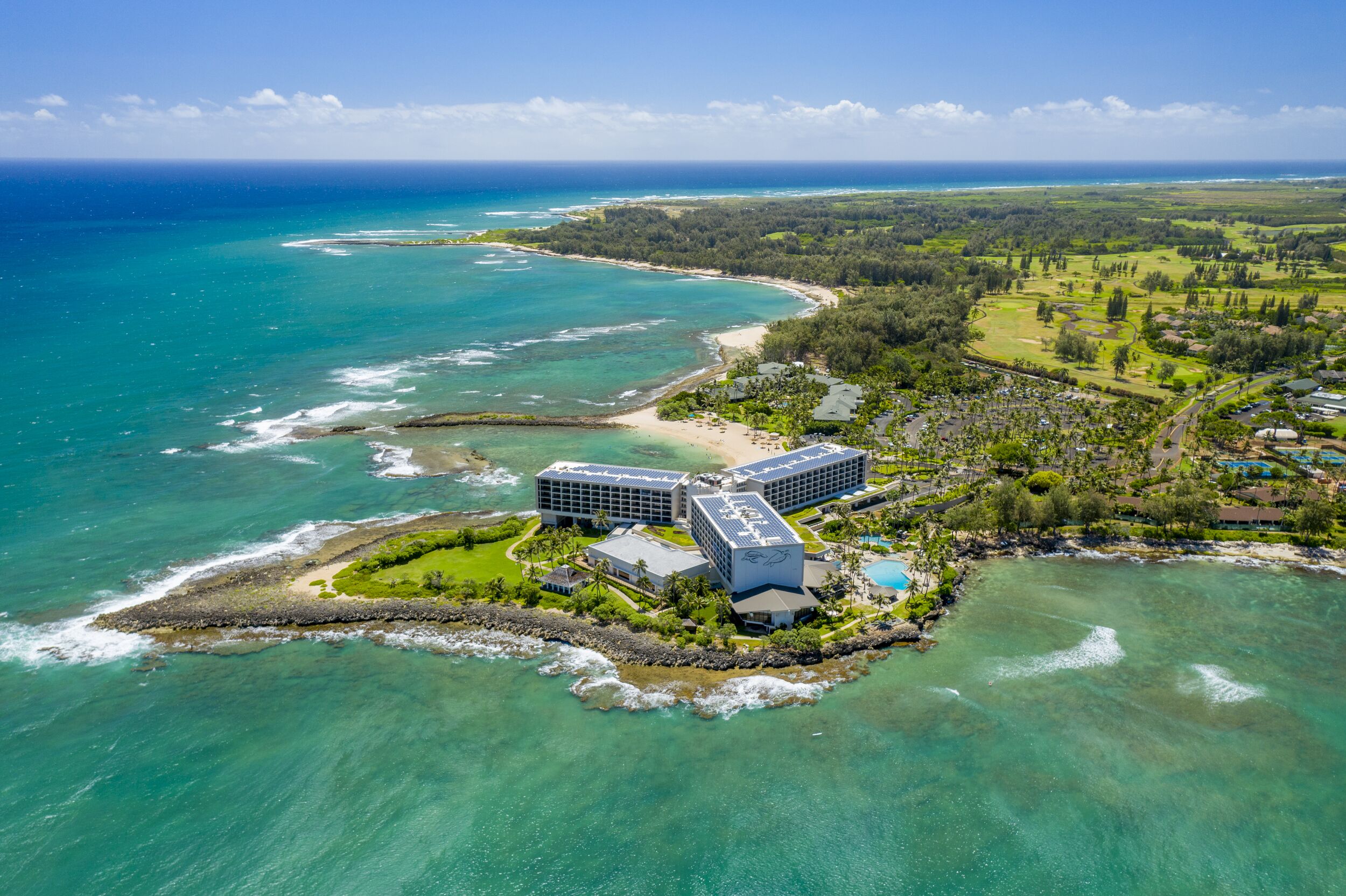 An aerial view of a coastal resort with a large hotel building, a beach, and clear turquoise waters.