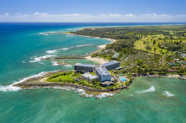 An aerial view of a coastal resort with a large hotel building, a beach, and clear turquoise waters.
