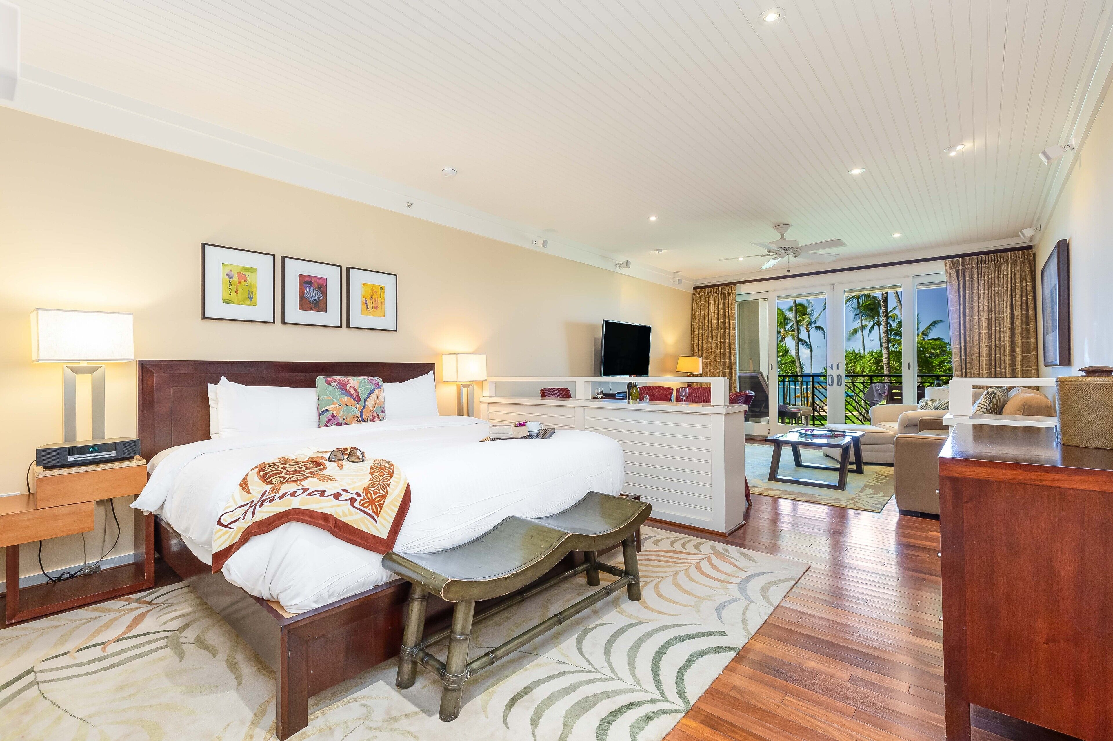 A hotel room with a large bed, bedside tables, a desk, a chair, a sofa, and a balcony with a view of palm trees.